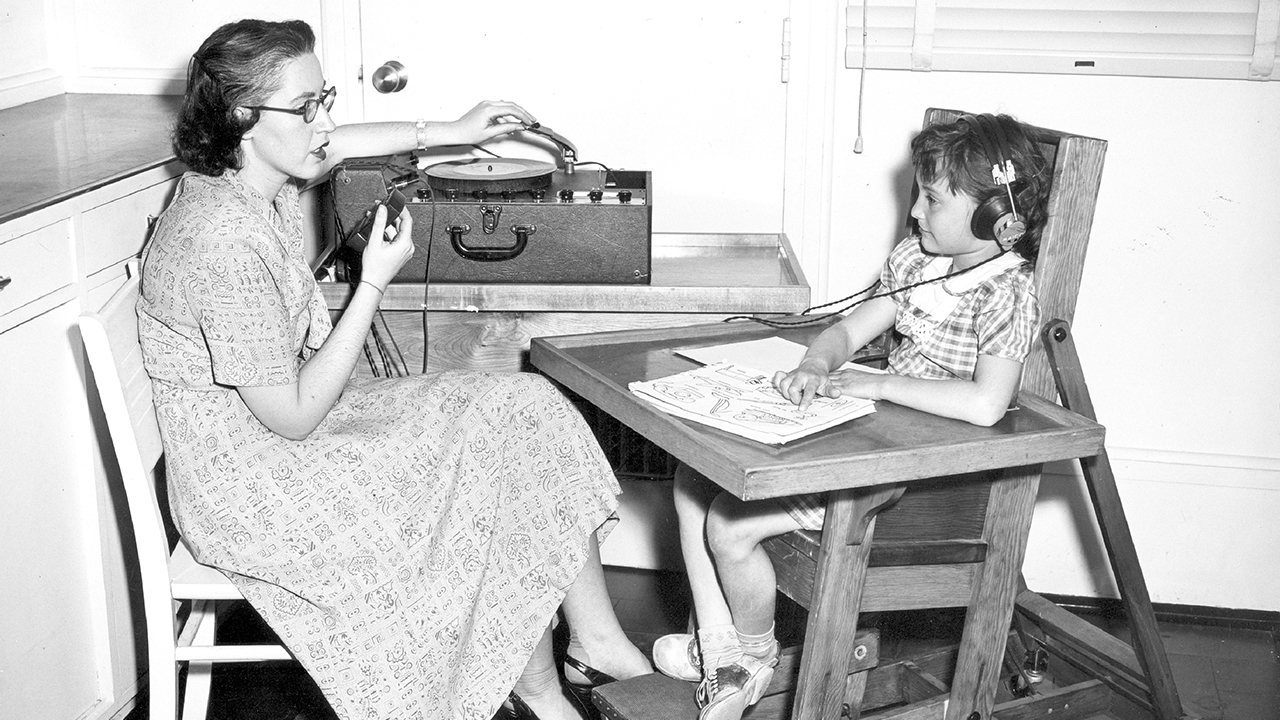 A black and white photo of a woman in a long dress with shoulder length brown hair and glasses speaking into a microphone and adjusting the speaker case while a young patient listens on headphones and sits in a chair with attached desk.