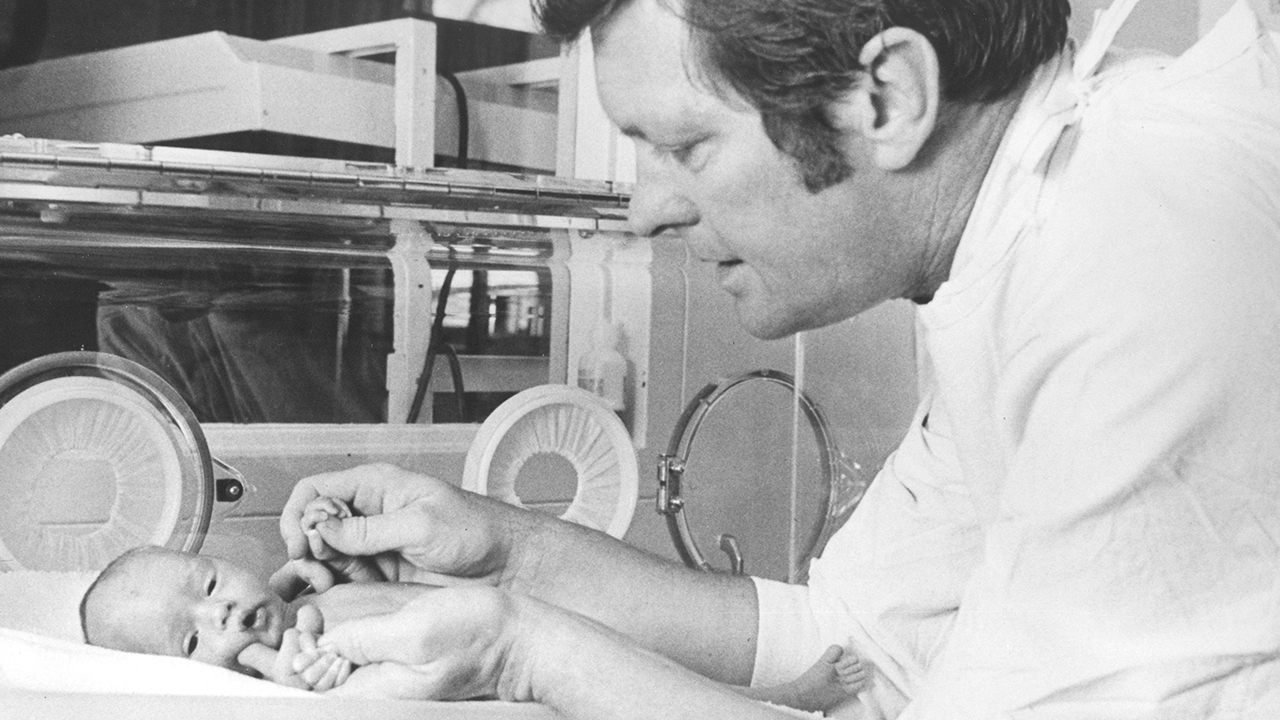 A black and white photo of Dr. Joseph Butterfield with dark hair and white shirt leaning over a newborn baby and holding its hands