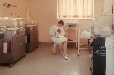 A nurse wearing a white dress and white hat sits in a rocking chair while holding a newborn baby. The room has newborn beds on each side and bright window on the back wall.