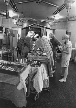 A black and white photo of an operating room with four surgeons working on a patient while a nurse looks on.
