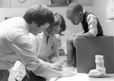 A black and white photo of Dr. Dennis Matthews with dark hair and a mustache standing next to a woman while they look at a young boy's foot. The boy is sitting on a small chair on the table and there is a foot brace sitting next to the chair.
