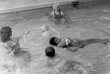 A black and white photo of two woman standing in a pool with balls and a hula hoop while two boys float in the water, one on his stomach with his head up and the other on his back wearing a life vest.