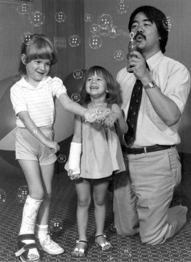 Black and white photo of Dr. Frank Chang with dark hair and a mustache, wearing a short sleeve shirt and tie, kneeling on the ground and blowing bubbles for two young girls trying to catch them. One girl has a cast on her foot and the other has a cast on her arm.