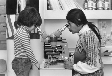 Black and white photo of a woman (Dr. Taru Hays) with long dark hair looking into a microscope with two viewfinders. A young kid wearing a striped shirt looks into the other viewfinder on the other side of the microscope.