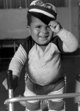 A black and white photo of a smiling toddler with braces on his legs and holding onto a walker with one hand and holding the brim of his spiderman hat with the other.