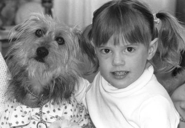 A black and white photo of a young girl with pigtails petting a small, shaggy dog