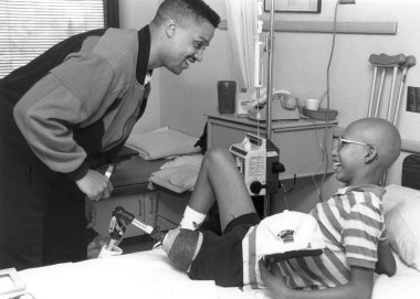 A black and white photo of Denver Nuggets basketball player Chris Jackson leaning over a patient and smiling while the boy looks up from the bed and smiles. The boy is holding a Denver Nuggets hat, wearing a striped shirt, has glasses and a prosthetic for his left leg.