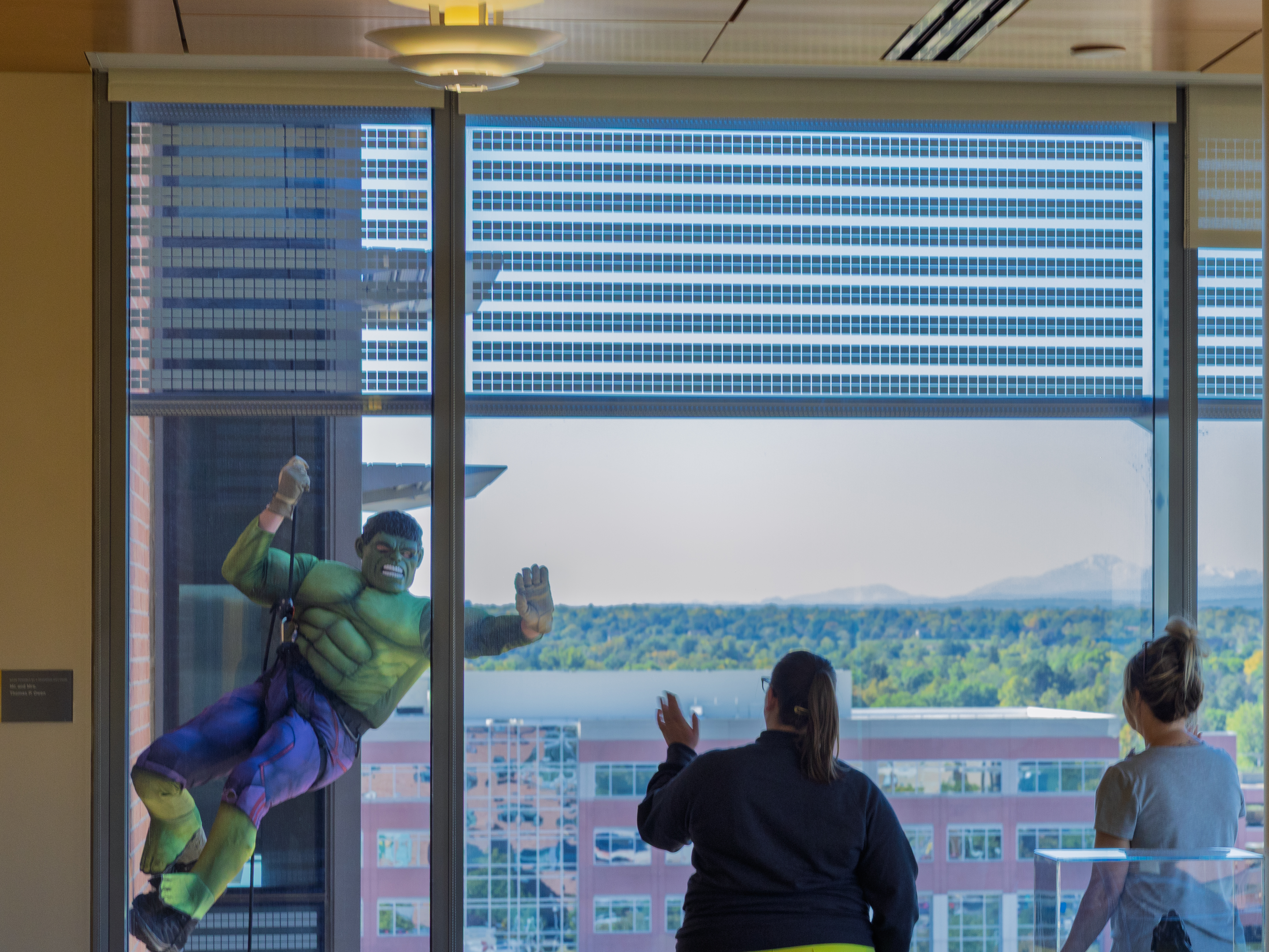 A law enforcement officer dressed as The Hulk superhero rappels down the side of Children's Hospital Colorado, Anschutz Medical Campus as part of a training exercise. The officer waves to two team members inside the building, who are waving back.