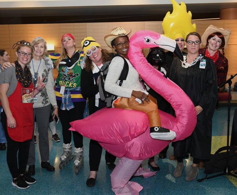 Group of people in costumes posing together indoors at an event, including an inflatable pink flamingo costume. 