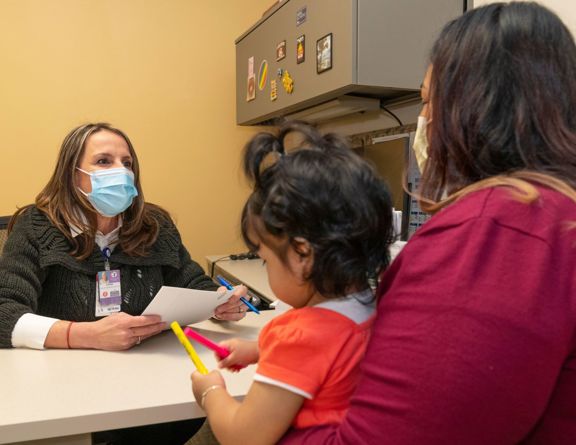 Person holding a child while talking to another person behind a desk while wearing masks 