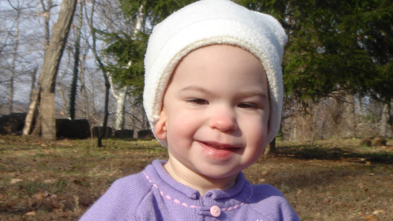 A toddler smiling, wearing a purple sweater and white snow hat