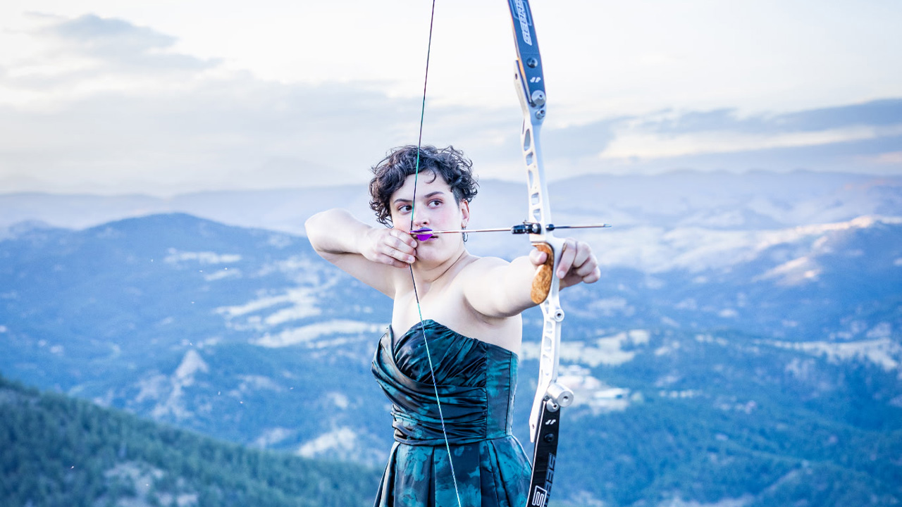A teenager with curly brown hair holding a bow and arrow, with mountains in the background
