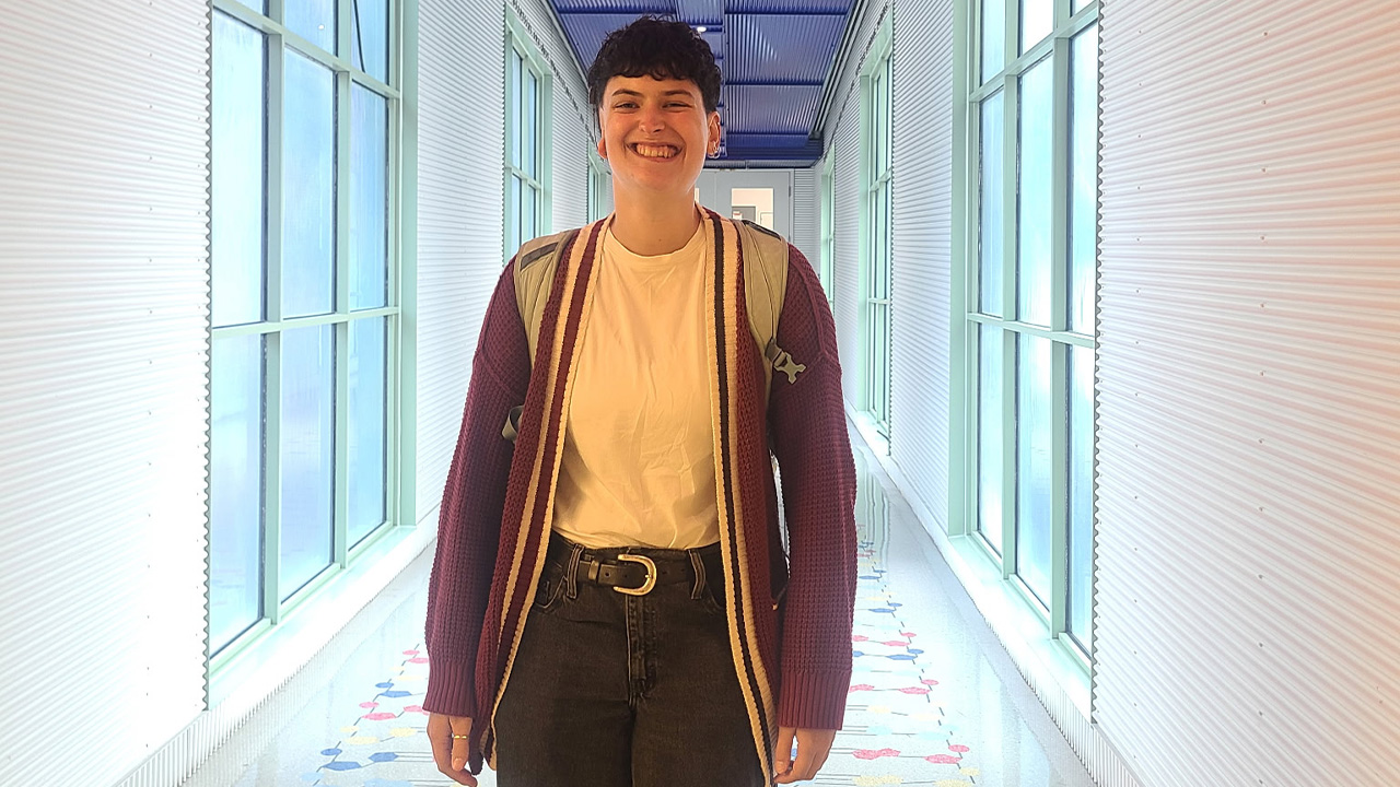 A teenager smiling wearing a red cardigan and a backpack, standing in a hallway lined with windows.