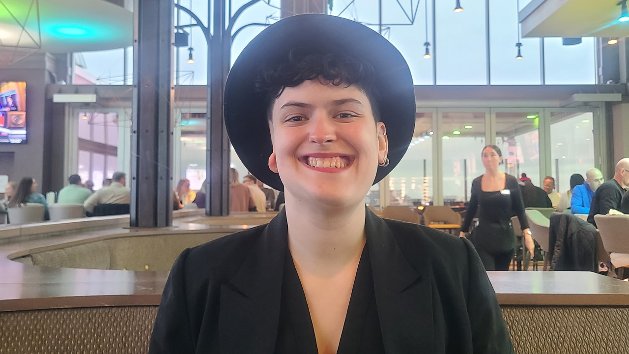 A teenager wearing a black brimmed hat and black jacket, smiling in a restaurant.