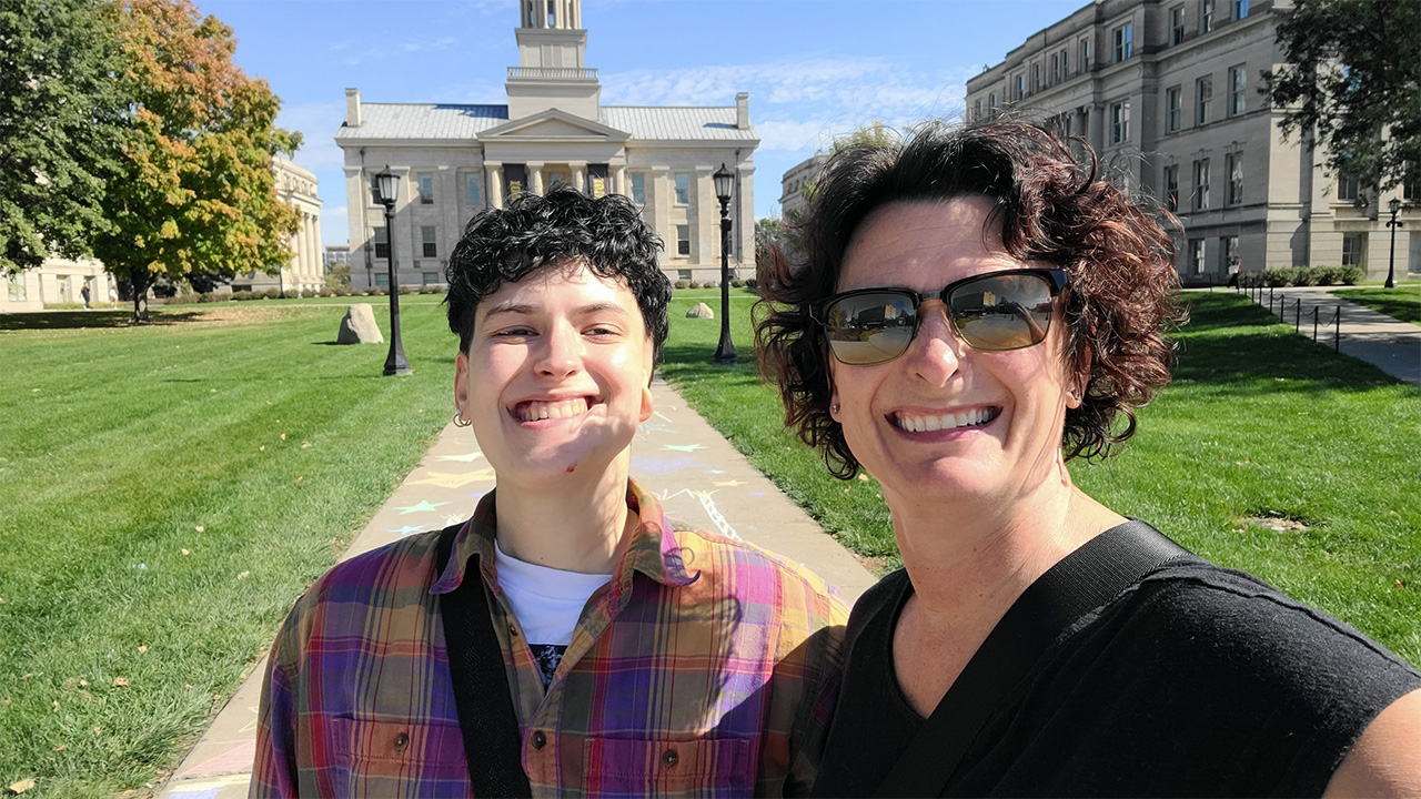 A young adult wearing a colorful flannel shirt and their parent standing outdoors on a sunny day, posing for a selfie in front of historic buildings.