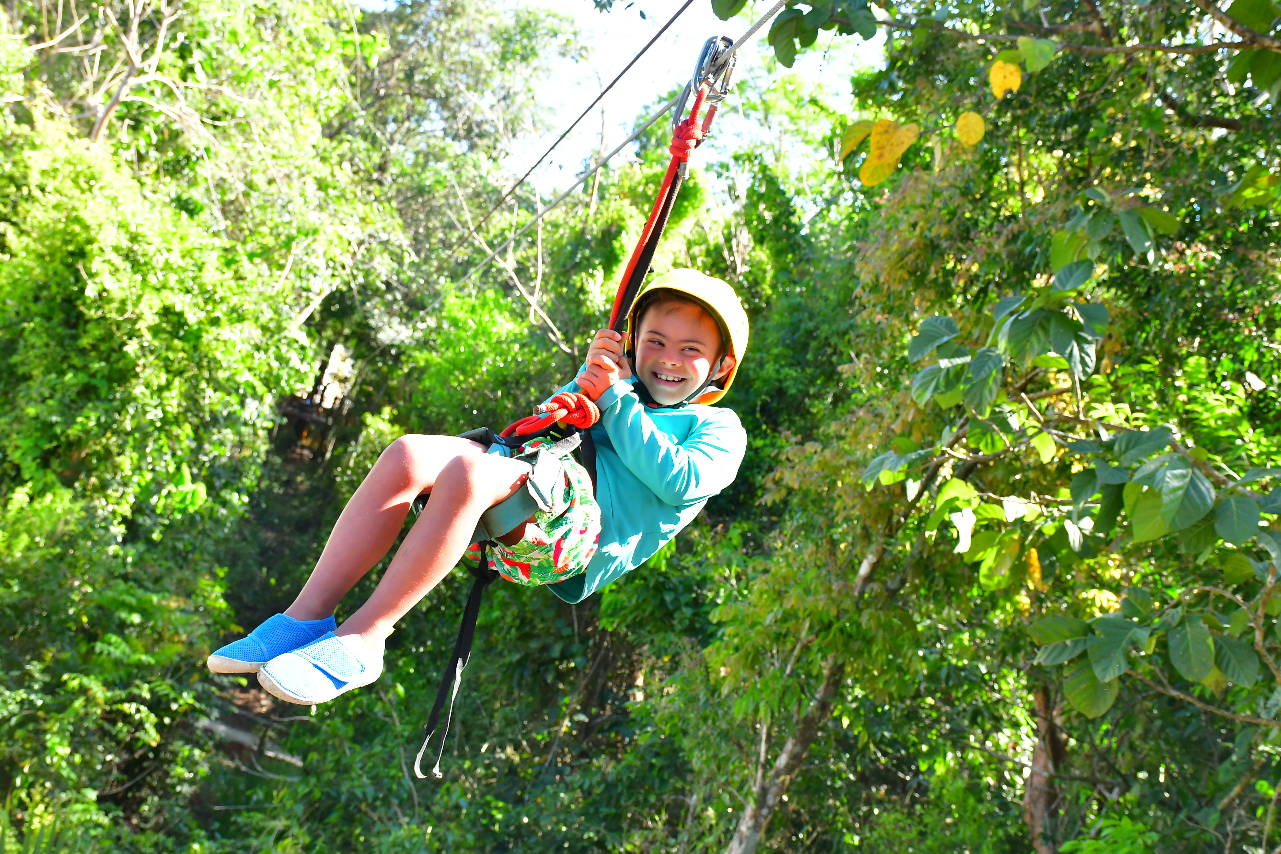 A child ziplines through trees wearing a harness and an orange helmet. 