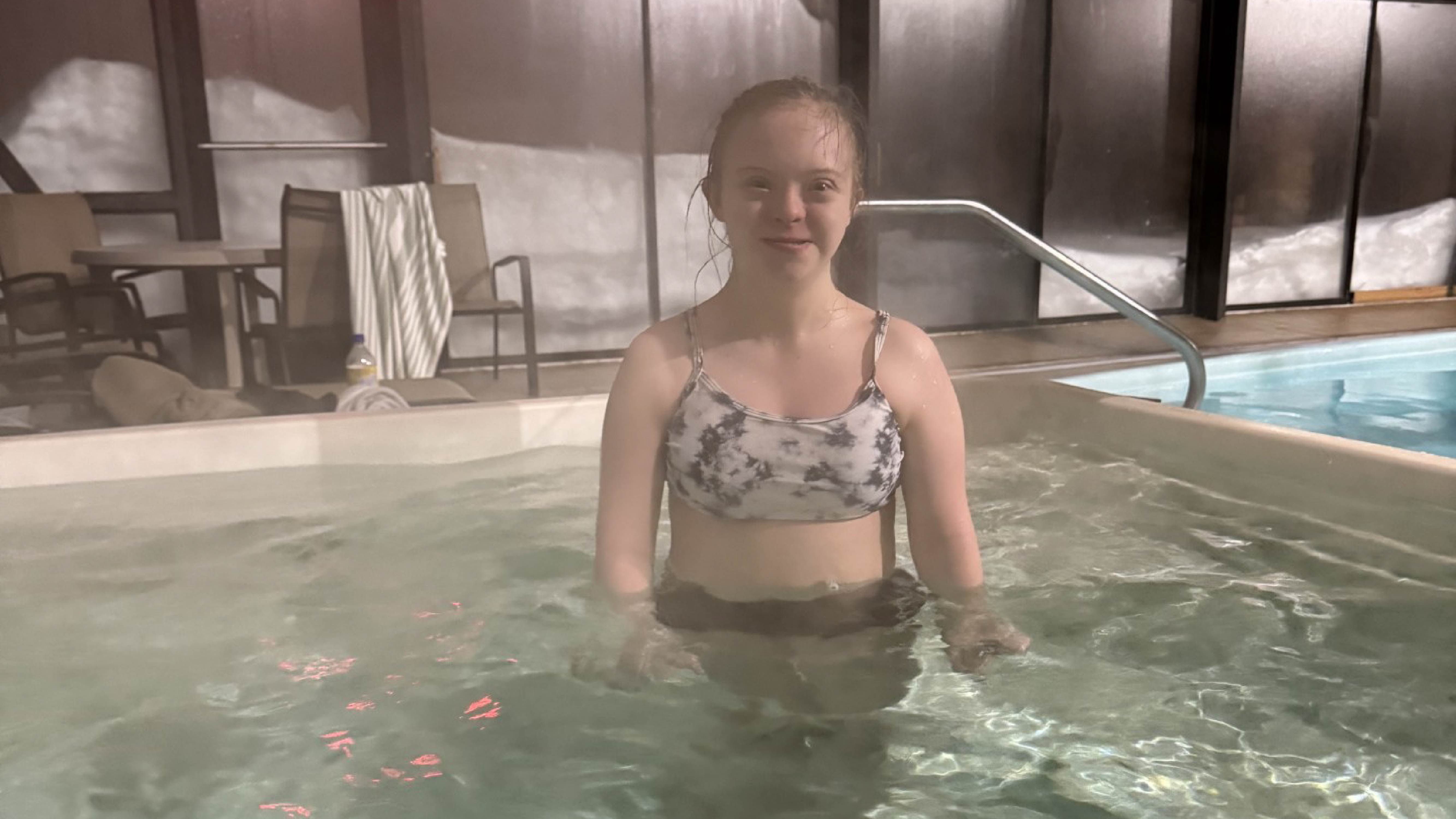 A young child stands in a hot tub while smiling.