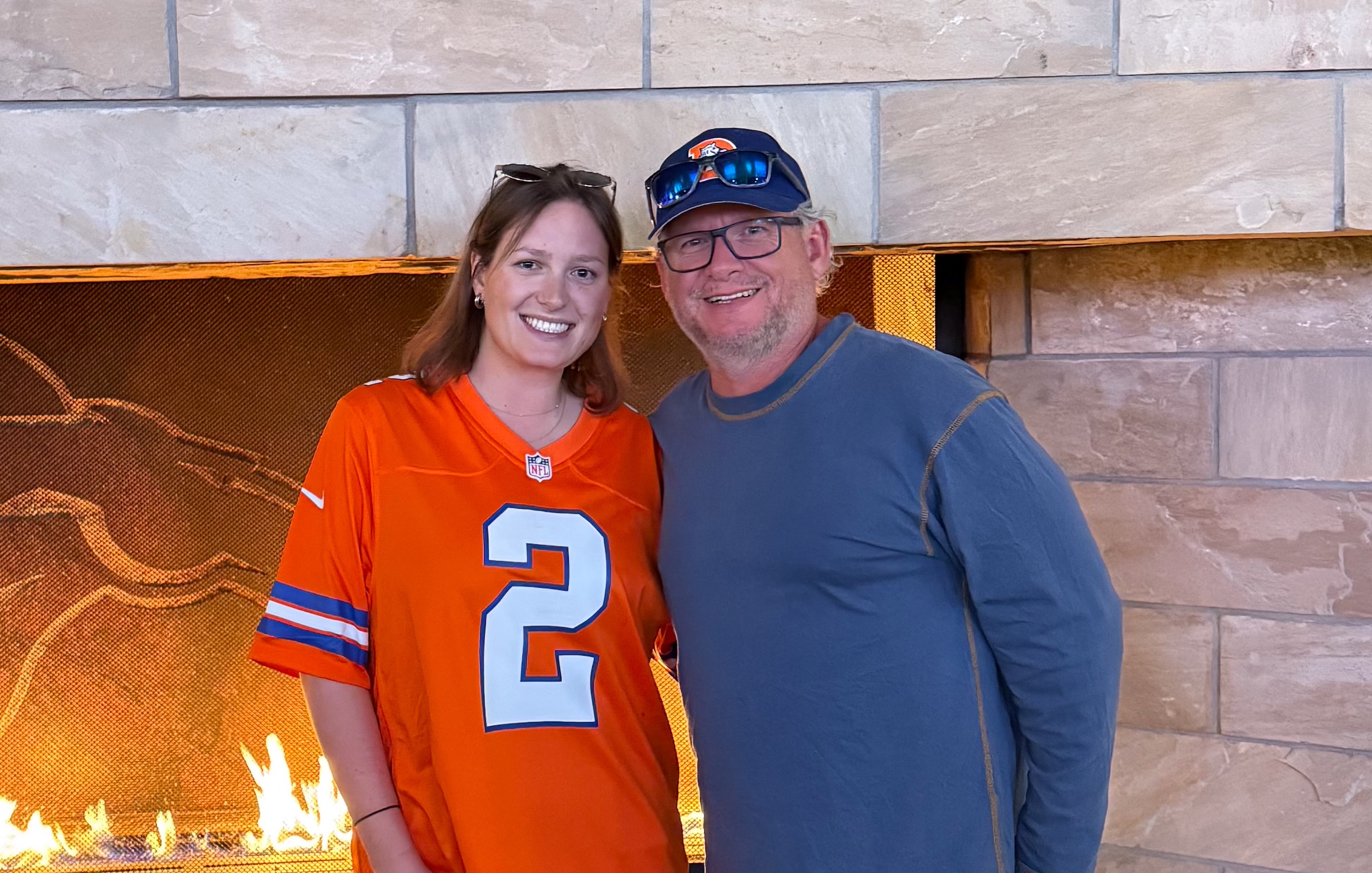 A woman in her 20s stands with her dad wearing Denver Broncos gear in front of a fireplace.