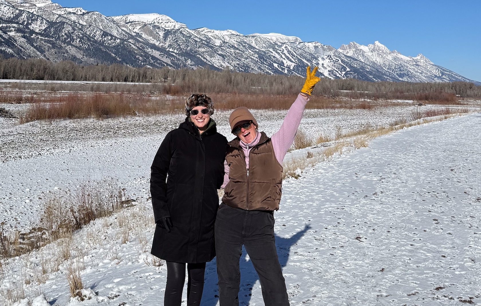 Two women stand together smiling in a snowy field in front of mountains. One holds her hand high in the air.