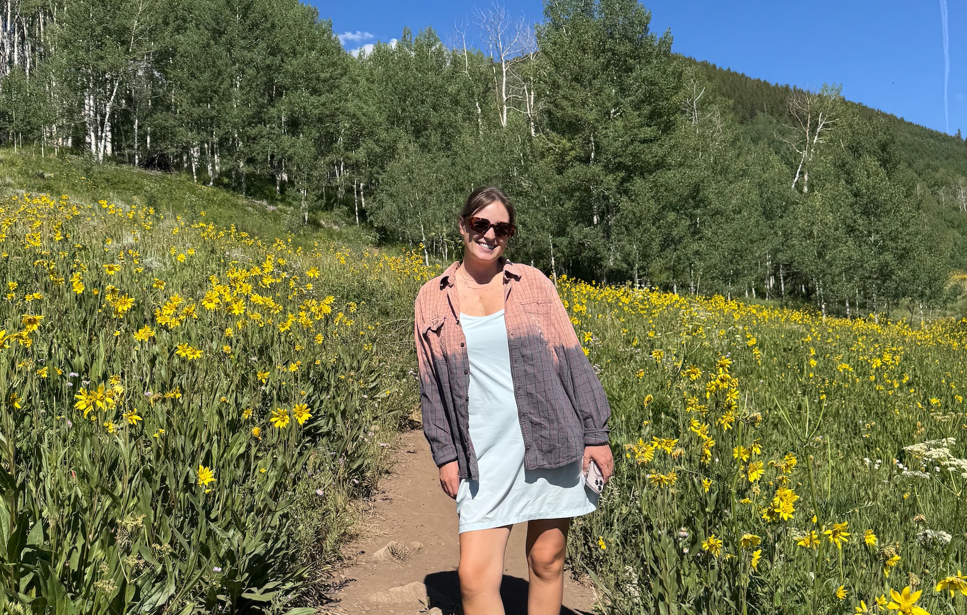 A woman in her 20s smiles while standing in a field of yellow wild-flowers in the mountains.