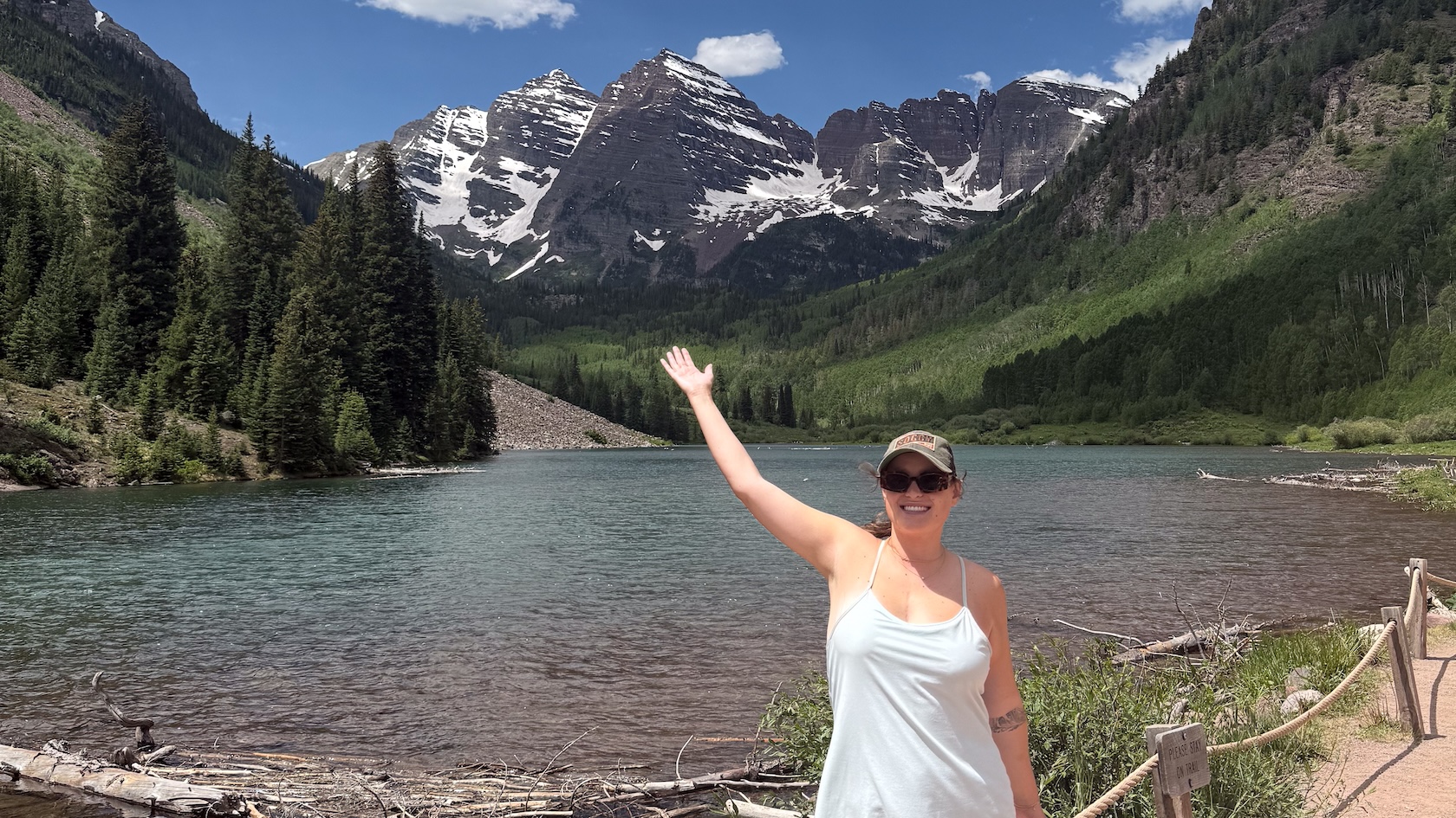 A woman in her 20s smiles and holds up her arm while hiking around a mountain lake.