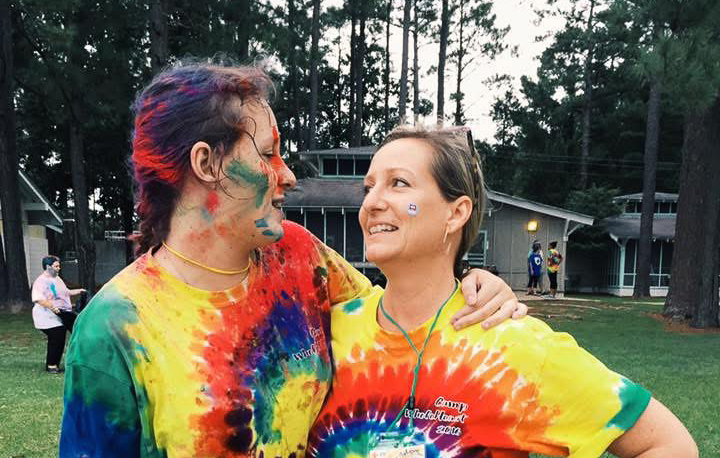 Two women stand together looking at each other while wearing tie-dye shirts. One has face and hair paint.