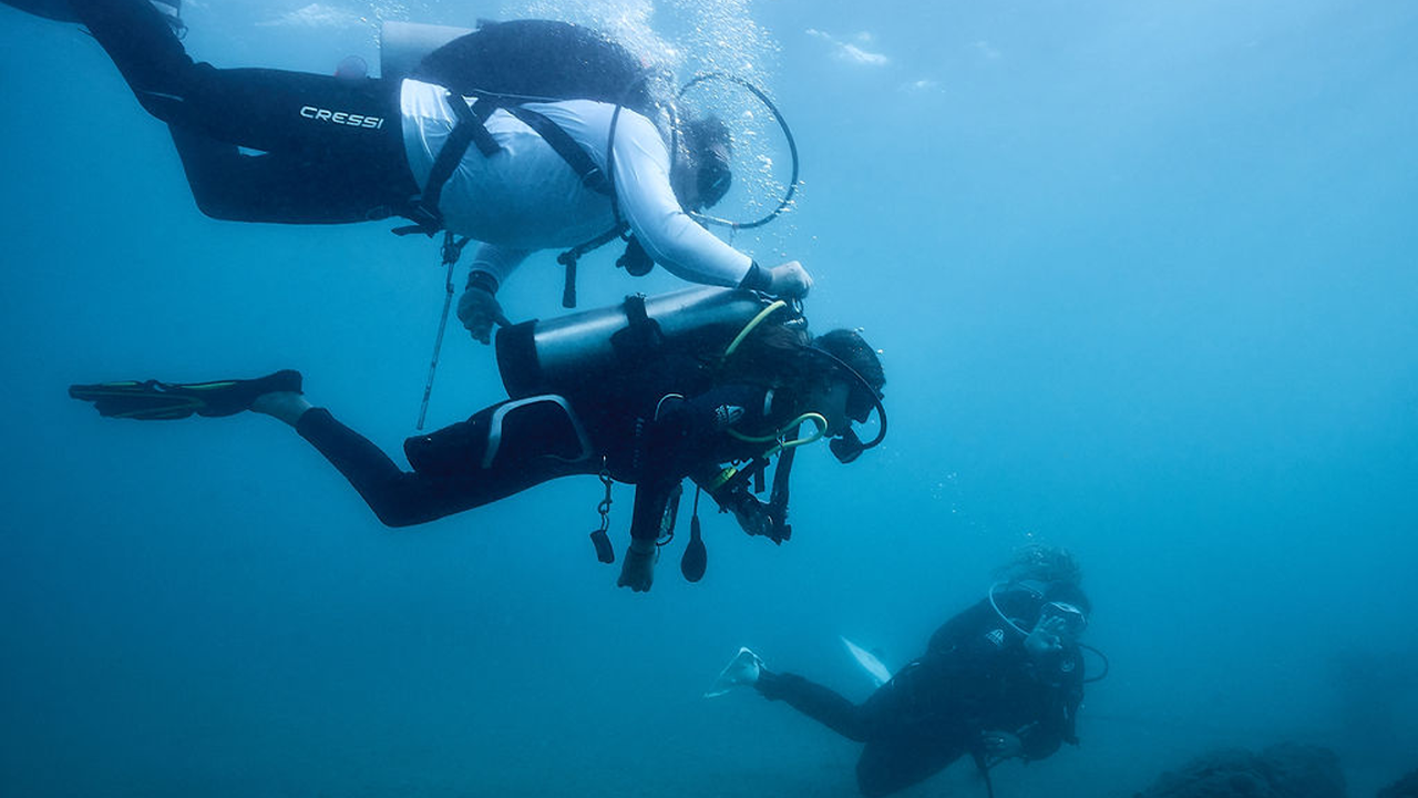 Two scuba divers in wetsuits and diving gear, including tanks, explore underwater near a rocky bottom.