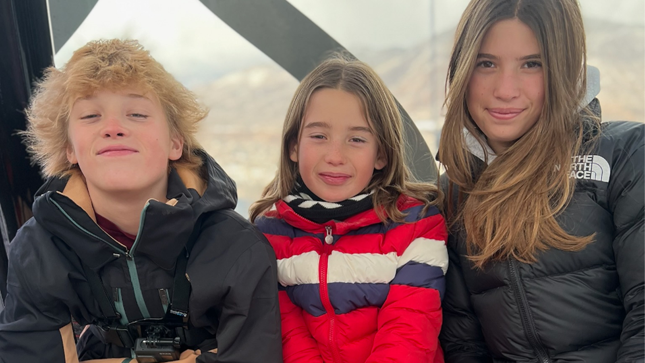 Three children smiling and sitting close together inside a ski lift gondola, dressed in winter sports gear.