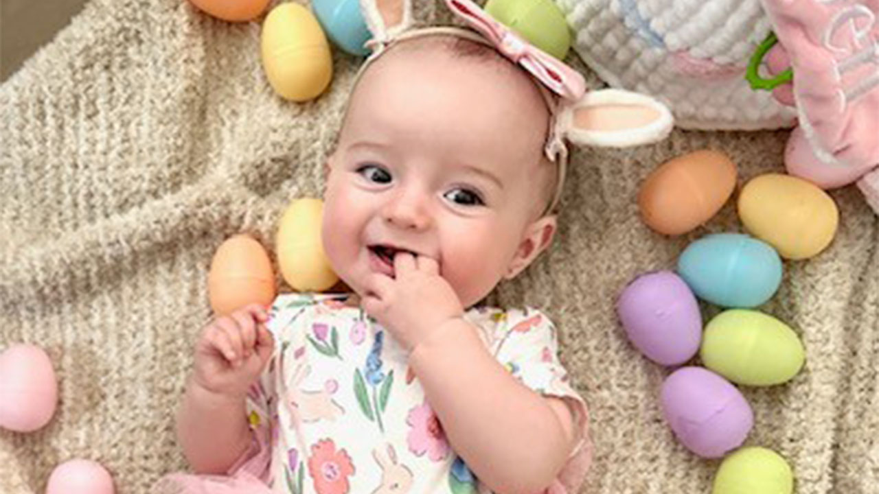 A baby with a flower shirt and pink bow lies amidst colorful Easter eggs while they gently hold two of their fingers in their mouth.