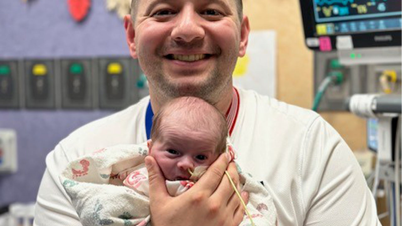 A parent smiles and holds their newborn baby in front of them on their chest while the baby has a nasal tube.