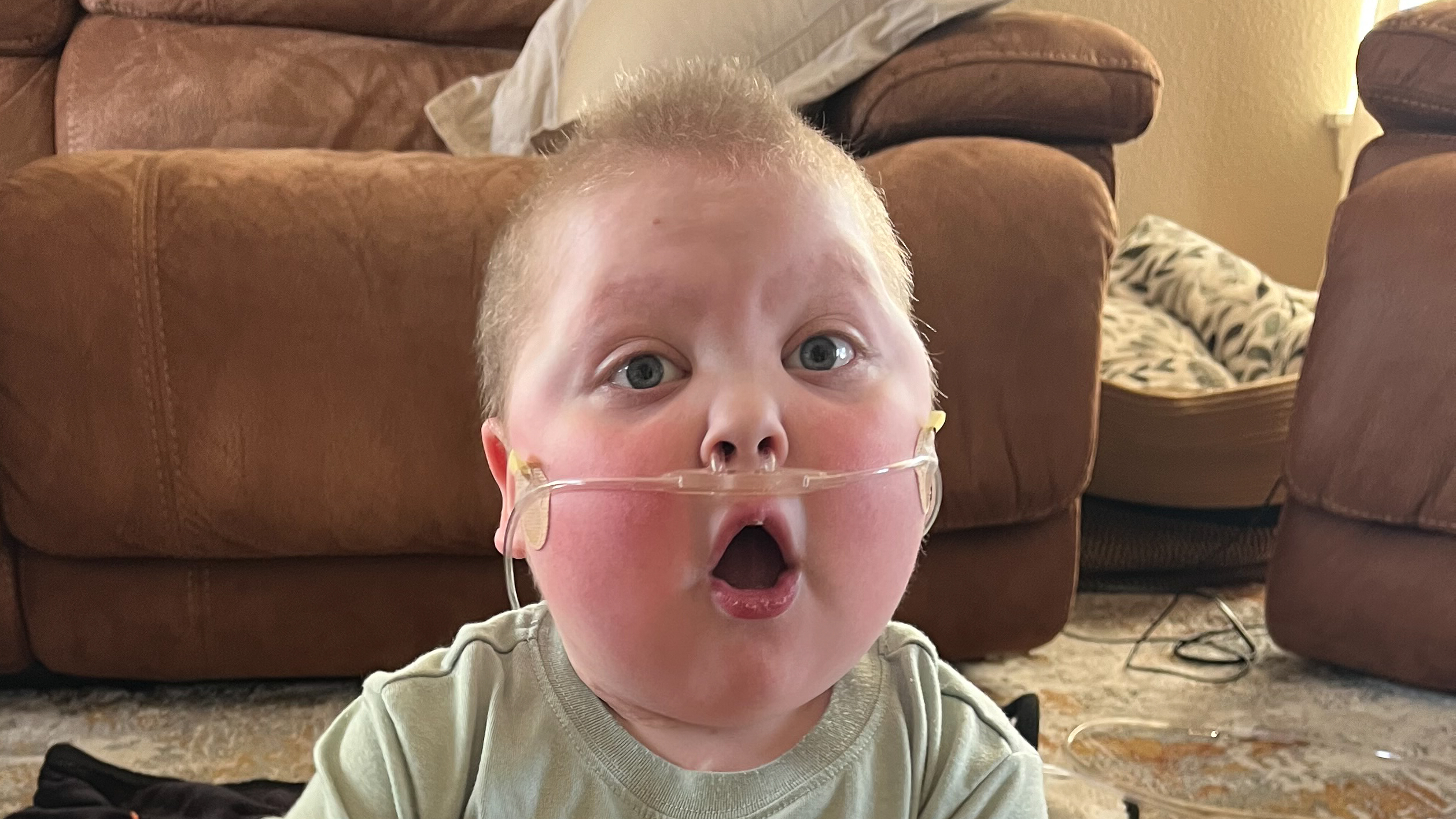 Conor is sitting indoors on a patterned rug in front of a brown upholstered couch showing a surprised face