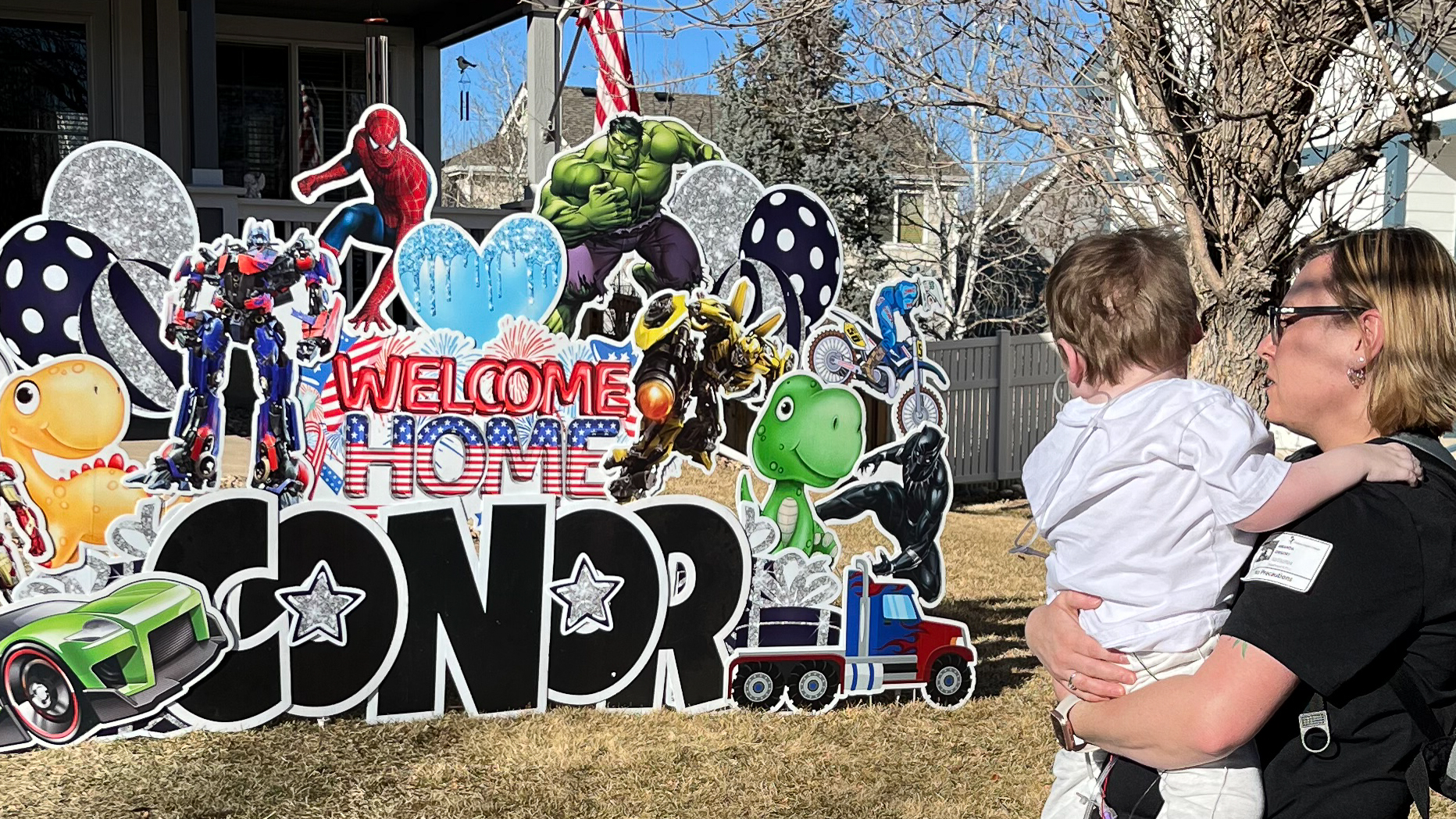 A colorful outdoor yard display reads 'WELCOME HOME CONNOR' in large letters surrounded by various cartoon and superhero characters. Conor’s mom is holding him while looking at the display on a sunny day in a residential neighborhood