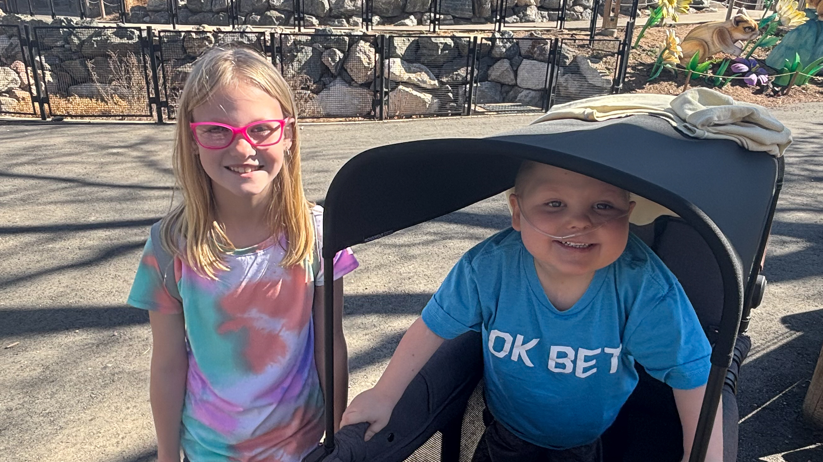 Conor and his sister, Brooklynn are outdoors on a sunny day. She is wearing a colorful tie-dye shirt, while Conor wearing a blue shirt with white text that reads 'OK BET.'