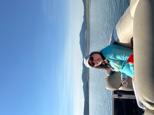 Person in a hat smiling on a boat, with clear blue skies and lake scenery in the background.