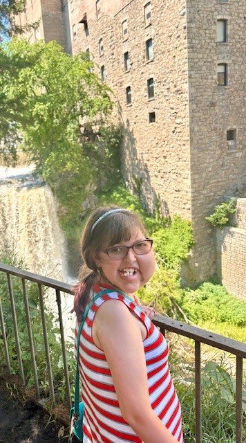 Person smiling at the camera with a scenic waterfall beside an old brick building in the background.