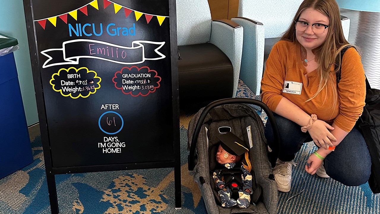 Katarina and Emilio pose next to a sign reading "NICU Grad." Emilio wears a small graduation cap.