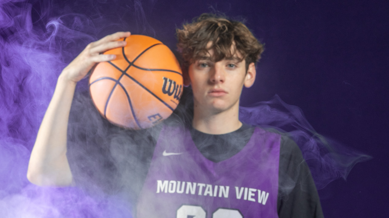 A person holding a basketball by their head with a purple jersey on and a black shirt underneath.