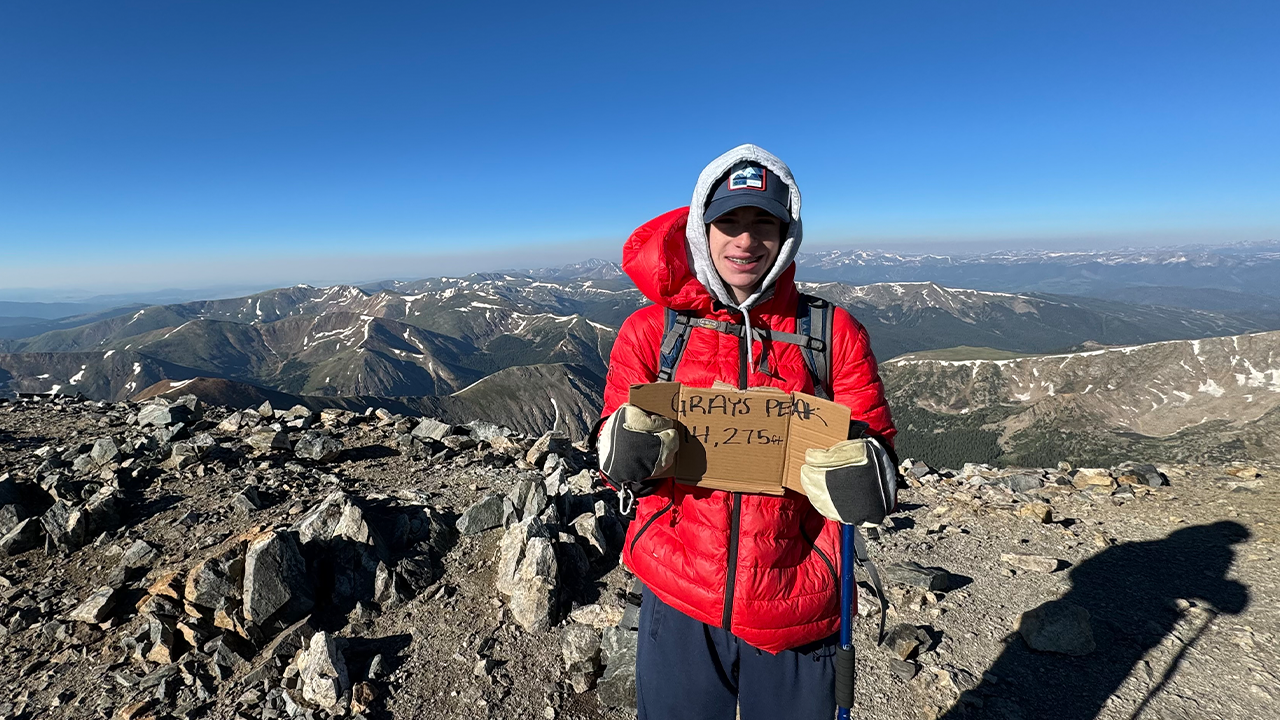 A person standing on top of a mountain wearing a red jacket and holding a sign of the hiking elevation.