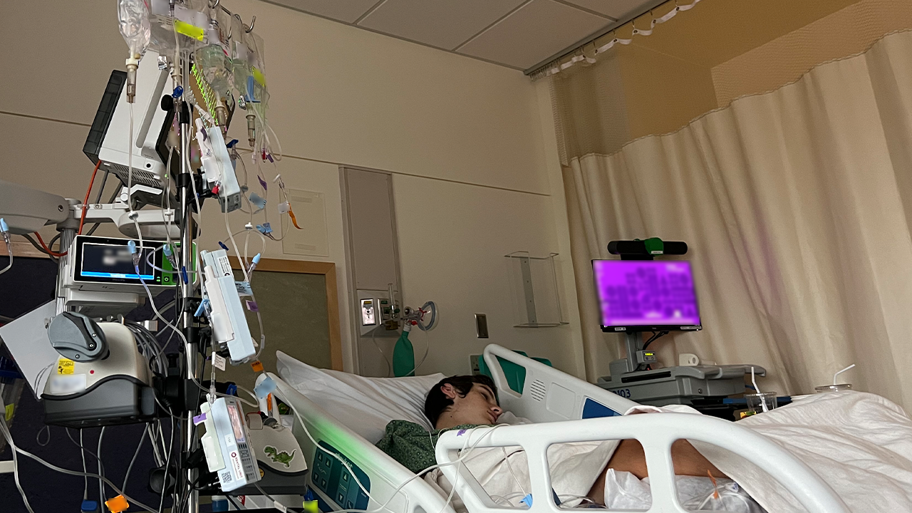 A person laying, sleeping, in a hospital bed with monitors and tubes surrounding them and a white blanket on top of them.