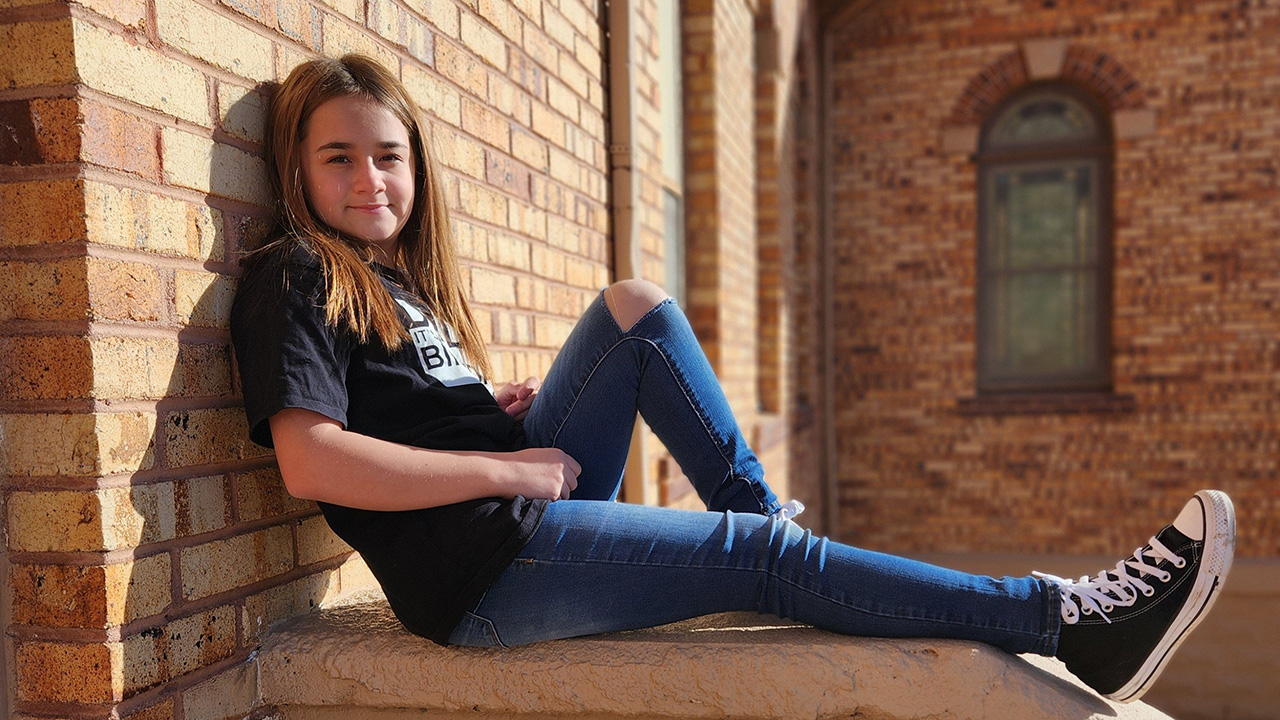 Jaycee sits against a brick wall in the sun and confidently smiles.