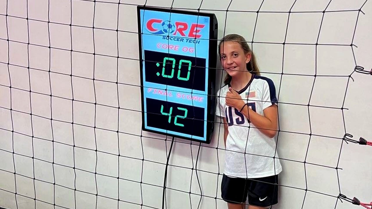 Kendall smiles with a thumbs up next to a soccer scoreboard.