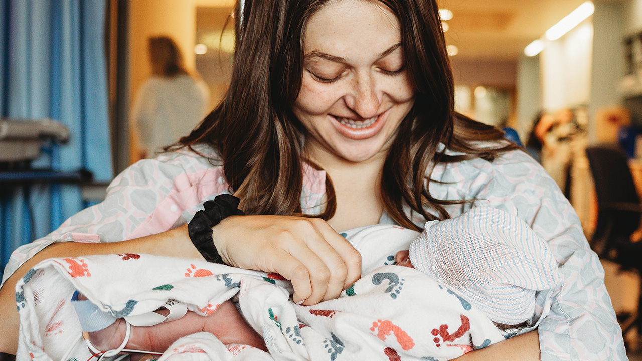 Mother smiling holding her newborn baby wrapped in hospital blanket with hospital hat on