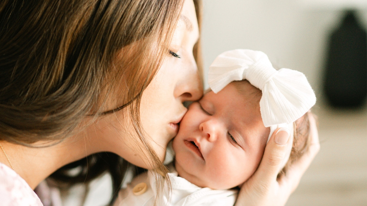 Mom kissing her newborn baby on the cheek. The baby wears a white bow on their head. 