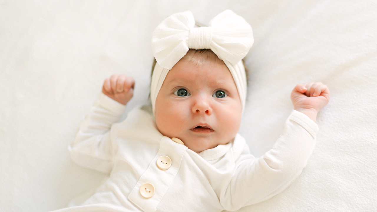 Newborn baby with a white outfit and white bow looking at camera