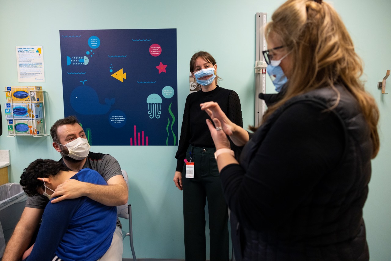 Juan watches a medical interpreter as Angel sits on his lap. There are clear plastic windows in the interpreter's masks for lip reading.