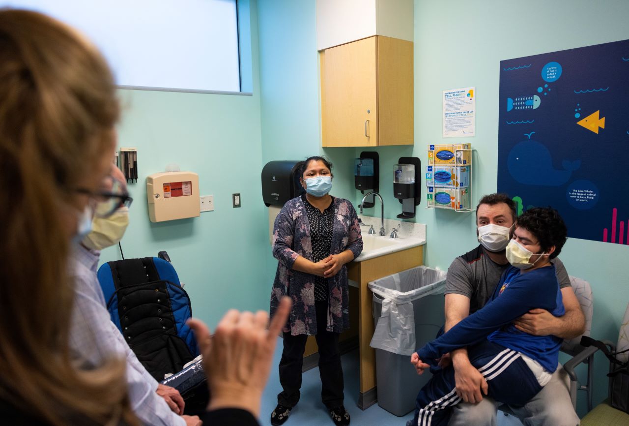 A medical interpreter speaks with Juan in sign language as Angel sits on his lap. Maria stands next to them as she listens to the providers speak.