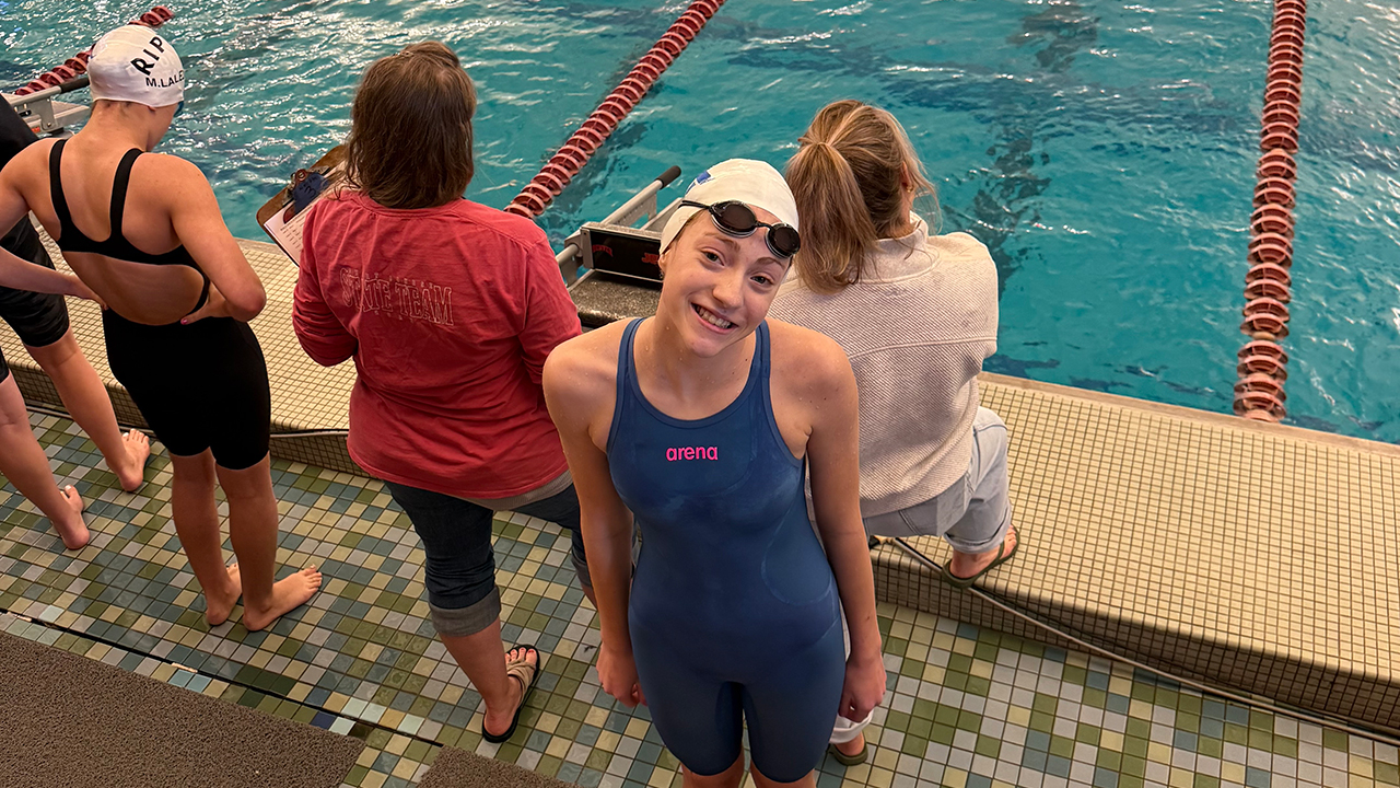 Young swimmer standing poolside at a swim meet with competition lanes visible in the background.
