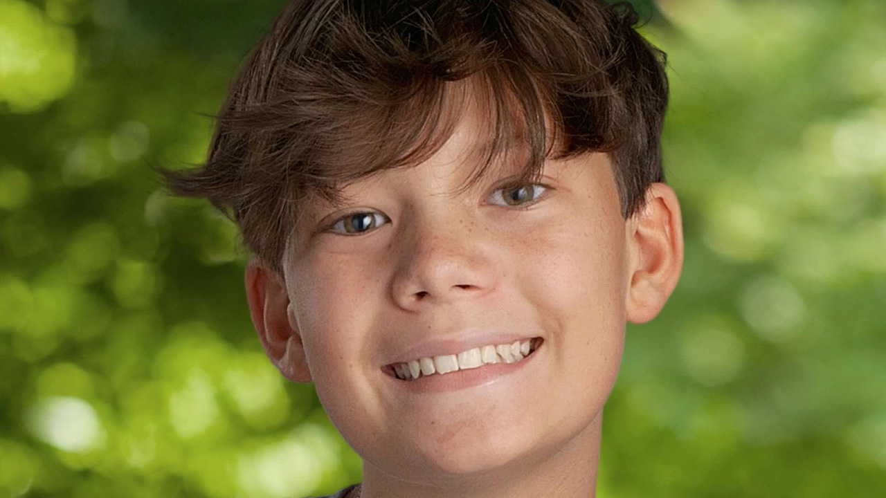 Portrait of a smiling young person wearing a grey t-shirt with a blue symbol, set against a blurred green foliage background.
