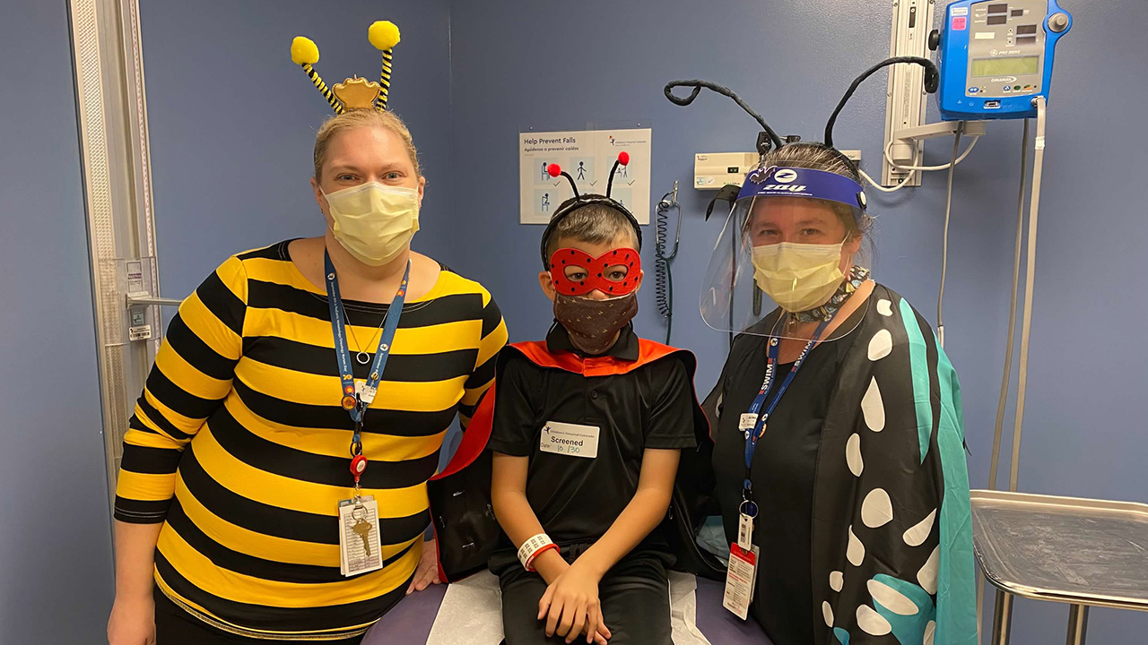 Three individuals in a medical room dressed in Halloween costumes: one as a bumblebee, another as a superhero and the third as a ladybug. All are wearing face masks and badges.