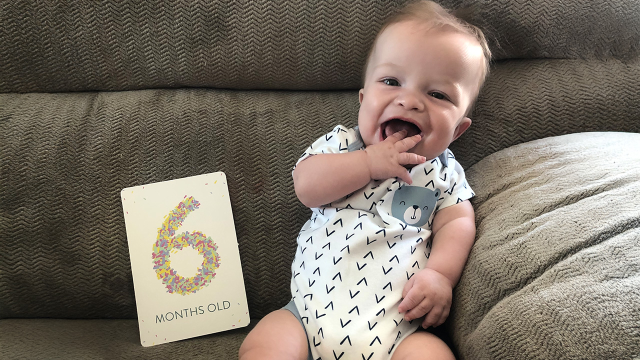 A baby sitting on a couch next to a sign that reads "6 months old," smiling and touching their mouth.